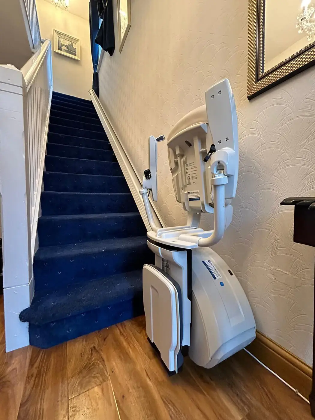 Straight stairlift with the seat folded at the bottom of a blue-carpeted staircase
